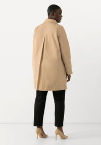 Trench coat beige, lunghezza al ginocchio, con una piega sul retro. Maniche lunghe e colletto, abbinato a pantaloni neri e tacchi alti beige.