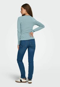 Langermet lys blå topp med bølget halskant. Kombinert med mørkeblå jeans og hvite sko med svart detalj. Glatt stofftekstur.