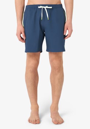 Costume da bagno da uomo blu navy con coulisse bianca e dettagli verde neon indossato da una persona in piedi a piedi nudi su sfondo bianco.