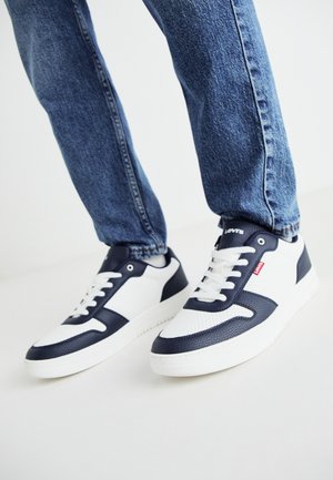 Zapatillas blancas y navy con detalles de cuero texturizado y malla, que cuentan con una suela blanca, cordones planos y una etiqueta de logo roja en el costado.