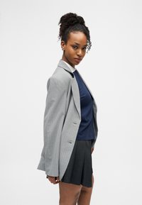 Νεαρή γυναίκα με σγουρά μαλλιά σε κότσο, που φορά γκρι σακάκι με λεπτές ρίγες, σκούρο μπλε πόλο πουκάμισο και μαύρη φούστα με πιέτες, στέκεται μπροστά σε λευκό φόντο.