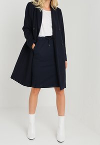 Manteau bleu marine à double boutonnage avec une silhouette ajustée, associé à une jupe côtelée assortie et des bottines blanches. Texture lisse tout au long.