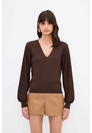 Brauner gestrickter Pullover mit V-Ausschnitt und strukturierten Puffärmeln; kombiniert mit hellbraunen Shorts, die den Kontrast in Farben und Materialien betonen.