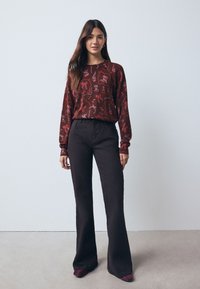 Haut à manches longues à motifs marron avec des dessins abstraits, associé à un pantalon noir évasé. La tenue présente une coupe décontractée et une texture lisse.