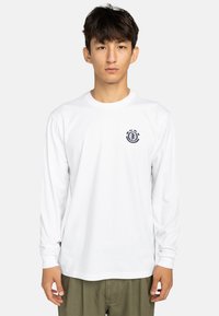 T-shirt bianco a maniche lunghe con scollatura rotonda e un emblema raffigurante un design ad albero in blu navy. Il tessuto appare liscio e leggero.