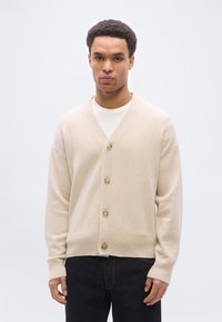 Νέος άνδρας που φοράει μπεζ κουμπωτή ζακέτα πάνω από λευκό πουκάμισο και σκούρο τζιν, στέκεται μπροστά σε απλό ανοιχτό γκρι φόντο.