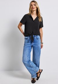 Zwarte kortebloes met een strik in de taille, gecombineerd met loszittende blauwe jeans en zwarte sandalen. Glad materiaal, klassieke kraag en casual pasvorm.
