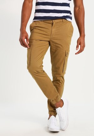 Homme portant un pantalon cargo marron avec des poignets élastiques, des baskets blanches et une chemise rayée bleu marine et blanche, debout avec une jambe croisée.