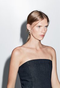 Šaty bez ramínek z tmavého denimu se strukturovaným korzetem, s hladkou texturou a čistými liniemi, doplněné náušnicemi ve tvaru kruhu.