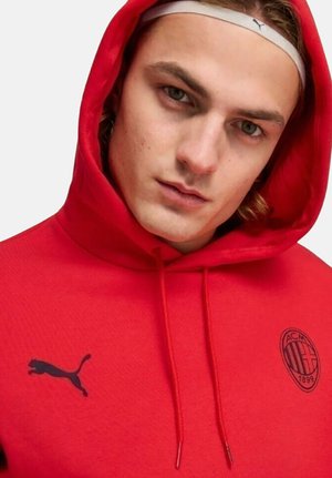 Giovane uomo che indossa una felpa rossa dell'AC Milan con il logo Puma e una fascia bianca Puma in testa, che guarda direttamente la fotocamera.