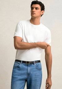Balta crew-neck marškinėliai, pagaminti iš medvilnės, derinami su mėlynais džinsais ir juodu diržu. Modelis stovi pasitikėdamas savimi, viena ranka sulenkta.