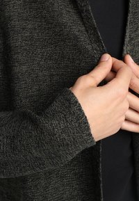 Mörkgrön stickad cardigan med ett strukturerat mönster, öppen front och rundade manschetter, vilket framhäver det mjuka, varma materialet.