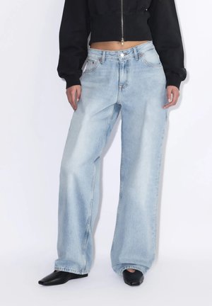 Persona che indossa jeans azzurri chiaro a gamba larga, scarpe nere con punta a mandorla e una felpa nera corta con zip.