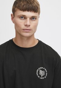 Sort bomulds t-shirt med et frontgrafikdesign, der har hvid tekst og blomsterillustration. Rund halsudskæring og korte ærmer.