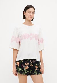 Witte t-shirt met korte mouwen, versierd met een roze bloemenprint op de borst, gecombineerd met zwarte shorts die bedekt zijn met kleurrijke tropische bloemenpatronen.