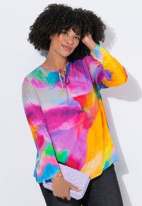 Vrouw met krullend haar draagt een kleurrijke blouse met aquarelpatroon, houdt een lavendelkleurige gewatteerde clutch vast en raakt haar haar aan tegen een witte achtergrond.
