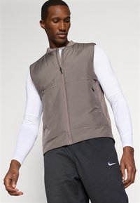 Gilet grigio in nylon con colletto rialzato e zip frontale. Presenta due tasche frontali e un piccolo logo sulla spalla sinistra. Indossato sopra una camicia a maniche lunghe.
