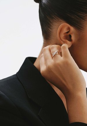 Primer plano de una mano con un anillo de oro apilado con pequeños diamantes tocando la oreja de una persona que lleva un blazer negro.