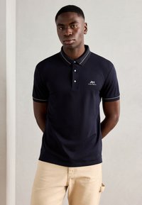 Marineblauwe polo met een kraag, een drieknoopsluiting en contrasterende lichtblauwe afwerking. Bevat een klein wit logo op de linkerkant van de borst.