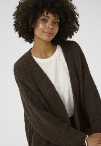 Brown knitted cardigan with a textured pattern, open front, and long sleeves, worn over a white t-shirt.