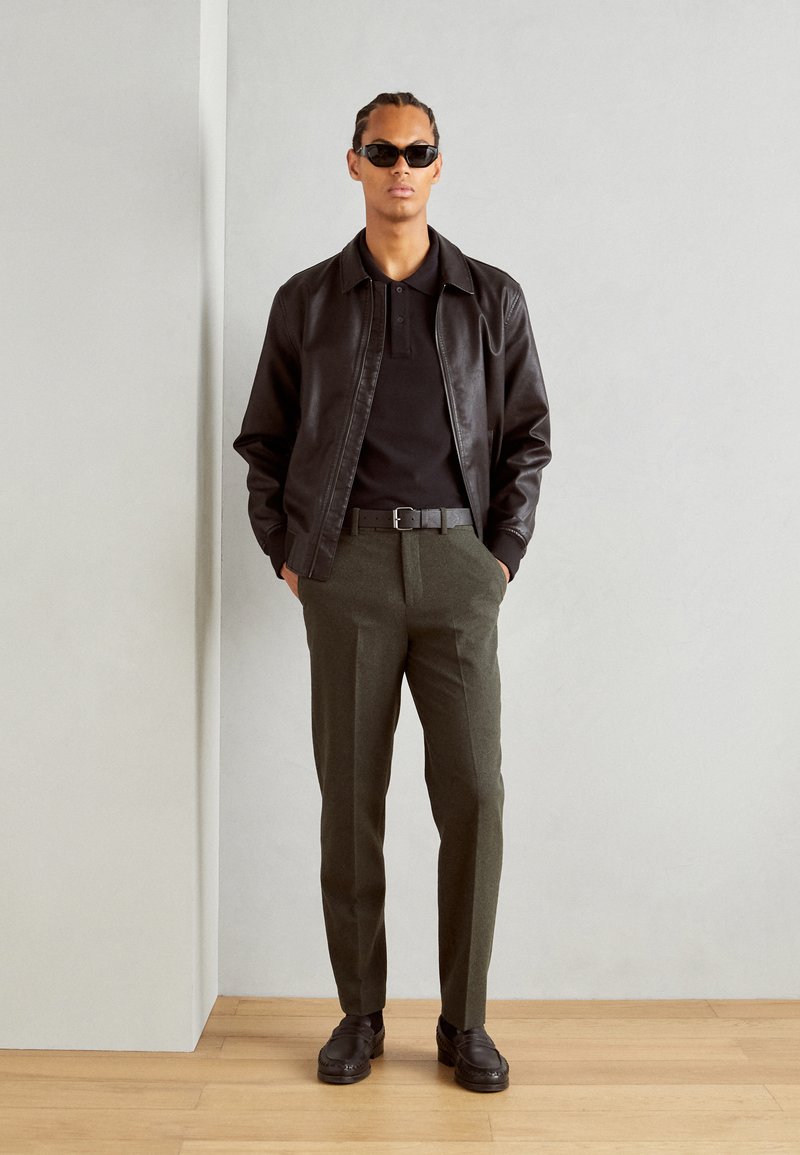 Schwarze Lederjacke über einem schwarzen Polohemd, olivgrüne Hosen, schwarzer Gürtel und braune Schuhe. Das Model trägt dunkle Sonnenbrillen.