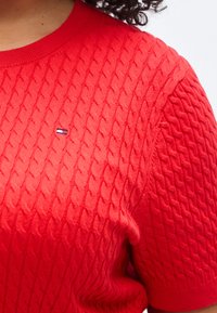 Κόκκινο πουλόβερ με κοντά μανίκια και πλέξη καλωδίου, με στρογγυλή λαιμόκοψη και μικρό λογότυπο σε μπλε και λευκό στο στήθος. Ορατή υφή υφάσματος.