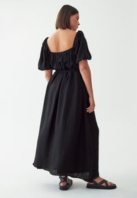 Black maxi dress with puffed sleeves, a deep square back, gathered waist, and soft texture. Paired with black sandals featuring a crossover strap design.