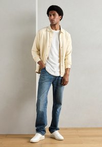 Giacca button-up gialla chiara sopra una maglietta bianca, abbinata a jeans blu e sneakers bianche. Tessuto morbido, vestibilità rilassata, stile casual.