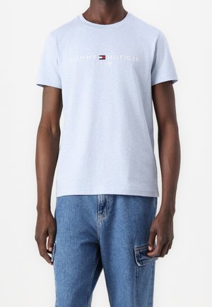 Mann trägt ein hellblaues Tommy Hilfiger T-Shirt und blaue Denim-Cargohose vor weißem Hintergrund.