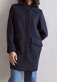 Mörk marinblå ullkappa med hög krage, två framfickor och rak passform. Vida ärmar och en ren, minimal design.