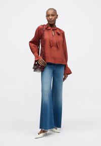 Blusa rossa con papillon e maniche lunghe, abbinata a jeans svasati blu chiaro. Una borsa trasparente bordeaux e mule bianche completano l'outfit.