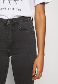 Jeans negros de cintura alta con una textura lisa, que presentan bolsillos frontales, un cierre de botón y costuras en contraste. Acompañados de una camiseta gráfica blanca.
