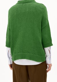 Pull en tricot vert avec une coupe ample, des manches courtes, un col et un ourlet côtelés. Porté par-dessus une chemise bleu clair et un pantalon marron.