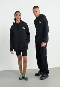 Black oversized hoodies with front pockets, neon green logo. Pairing with black cycle shorts and contrasting footwear with bright laces.