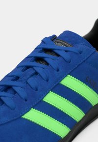 Μπλε αθλητικό παπούτσι Adidas Gazelle με φθορίζουσες πράσινες ρίγες και κορδόνια στο ίδιο μπλε χρώμα, πάνω σε λευκό φόντο.