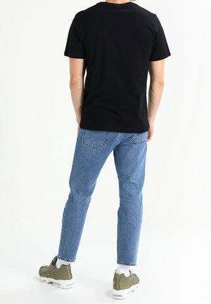 T-shirt en coton noir associé à un jean bleu clair et des baskets vert olive, vue de dos, avec des coutures et des poches visibles.