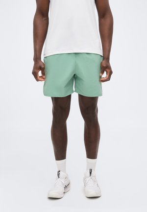 Uomo con pantaloncini sportivi verde chiaro, calzini bianchi, scarpe da ginnastica bianche e una maglietta bianca, in piedi davanti a uno sfondo chiaro uniforme.