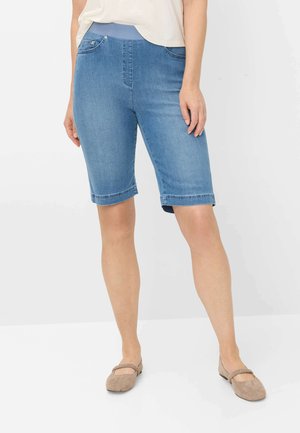 Vrouw draagt knielange denimshorts met elastische tailleband en beige platte schoenen, staand tegen een eenvoudige lichte achtergrond.