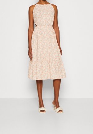 Femme portant une robe midi sans manches, couleur pêche claire à motif floral avec des découpes sur les côtés, associée à des sandales tressées à talons hauts couleur crème.