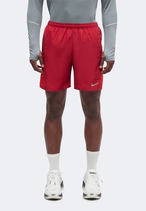Homme portant un short de sport Nike rouge, un t-shirt gris à manches longues, des chaussettes blanches et des chaussures de course Nike blanches, debout devant un fond uni.
