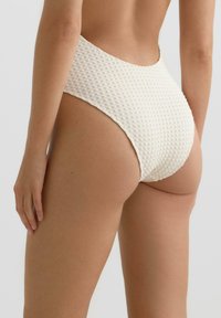 Žena v bielych bikínach s vysokým pásom a textúrou, zobrazená zozadu na neutrálnom svetlom pozadí.