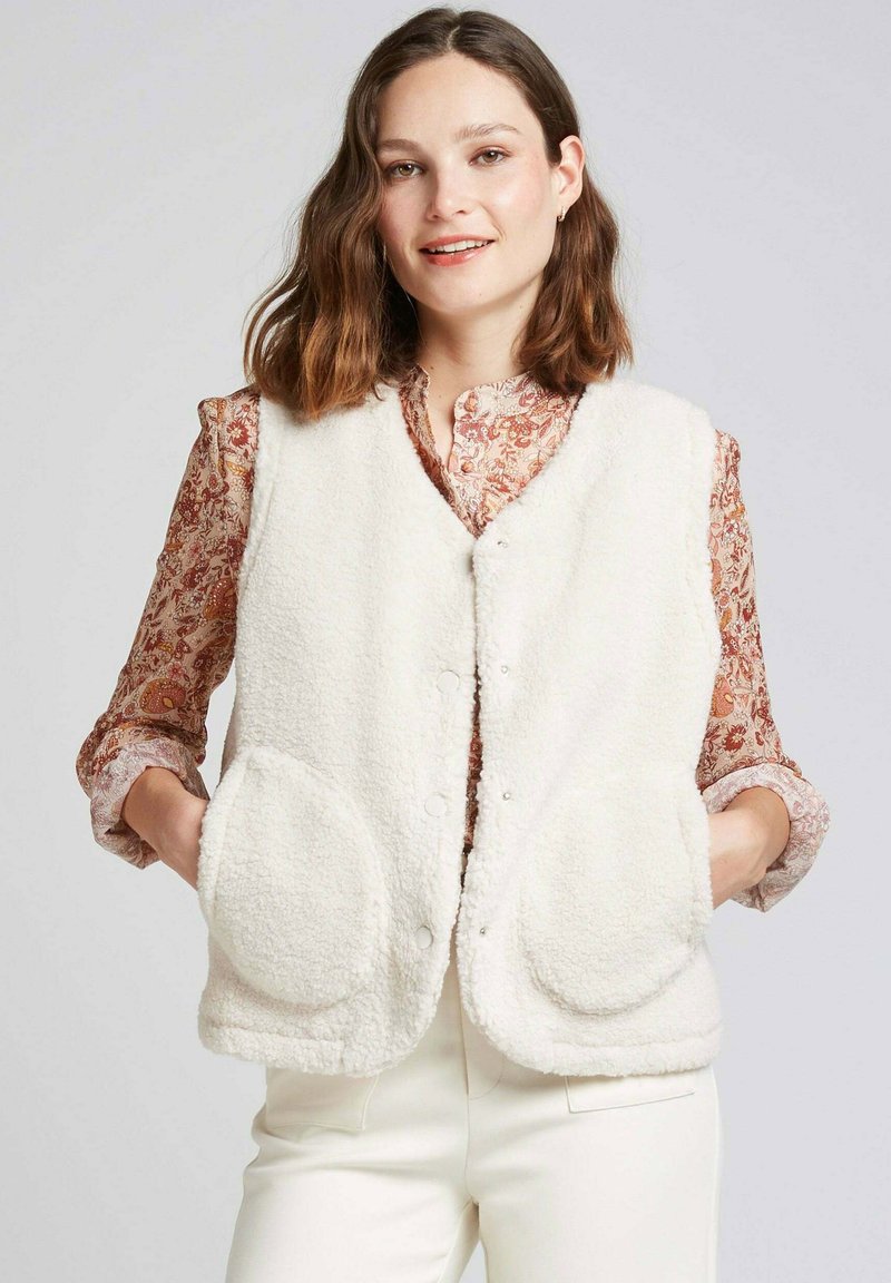 Gilet en polaire blanc avec un col en V, fermeture à pression à l'avant, et deux grandes poches rondes. Porté par-dessus une blouse à motifs.