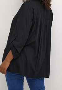 Svart långärmad skjorta i lätt tyg, med veckad bakdel, uppvikta ärmar och lös passform, ihop med blå jeans.