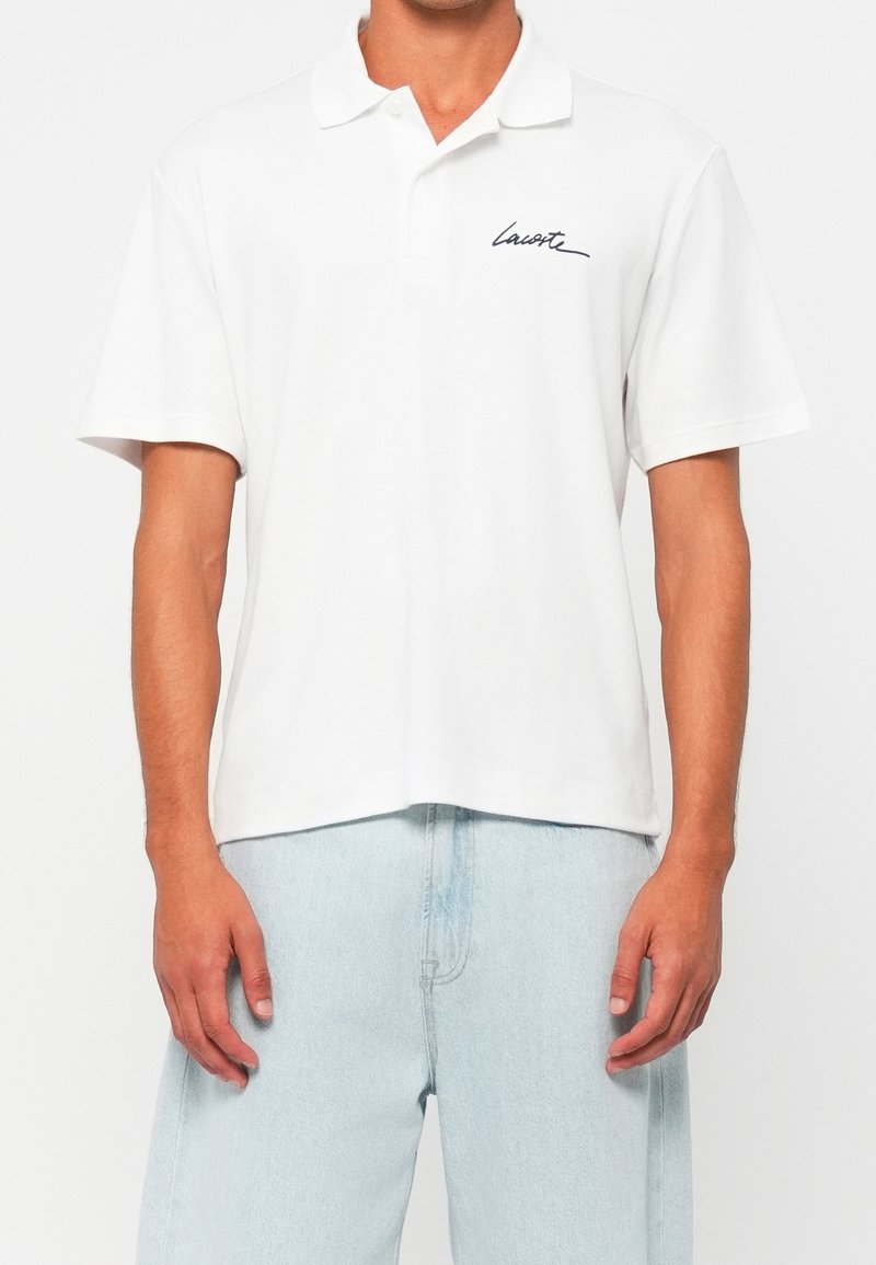 Homme portant un polo blanc à manches courtes avec le logo "Lacoste" sur la poitrine et un jean bleu clair, de face sur fond uni.
