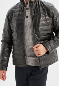 Giacca imbottita nera con finitura testurizzata, colletto alto, chiusura con cerniera e linee cucite. Indossata sopra un maglione grigio chiaro e pantaloni blu.