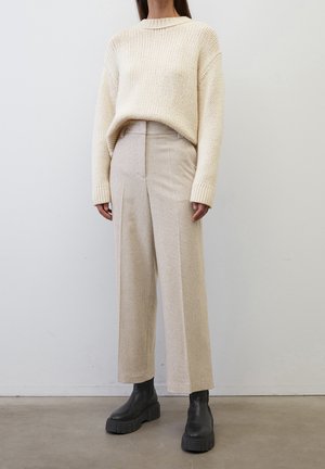 Nainen pukeutuneena vaaleaan neulepaitaan, beigenvärisiin korkeavyötäröisiin lyhytlahkeisiin housuihin ja mustiin paksupohjaisiin saappaisiin, seisomassa valkoisen seinän edessä.