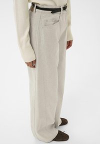 Persona, kas ģērbjas krēmkrāsas plašās biksēs, ar melnu jostu, krēmkrāsas garajās piedurknēs džemperī un tumši brūnās mīkstajās kurpēs, stāv sānis.