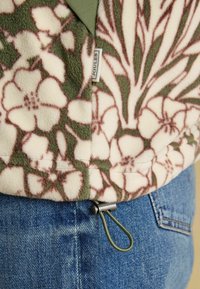 Fleecová bunda s květinovým vzorem v zelené a krémové barvě. Obsahuje štítek a nastavitelné šňůrky na spodním lemu, kombinována s modrými džíny.