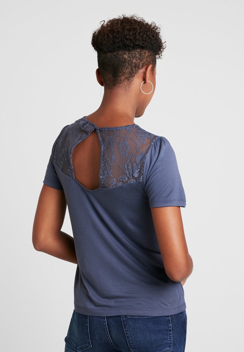 Personne aux cheveux courts et bouclés portant un haut bleu à manches courtes avec de la dentelle fleurie et un détail œillet dans le haut du dos, associé à un jean bleu.