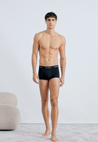 Crne pamučne bokserice s Calvin Klein logotipom na pojasu, koje nosi goli muški model na svijetloj pozadini.